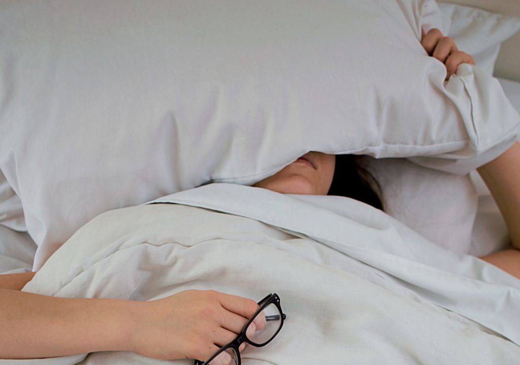 person lying on bed while covering face with pillow and holding eyeglasses
