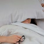 person lying on bed while covering face with pillow and holding eyeglasses